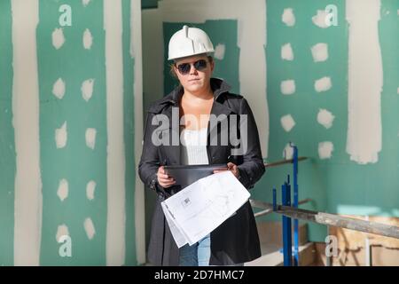 Young architect wearing a safety helmet and sunglasses standing at a construction site and looking at the camera while holding the paper floor plans Stock Photo