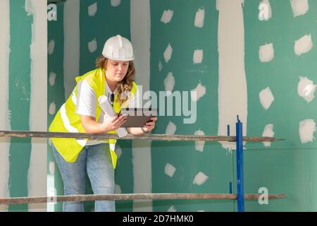 Architect wearing white helmet and a safety jacket leaning against a scaffolding and checking out the floor plans on an electronic device Stock Photo
