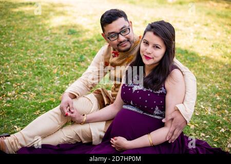 Young asian indian pregnant woman with her husband in sitting relaxing in park or garden, young parents looking at camera expecting baby. Stock Photo