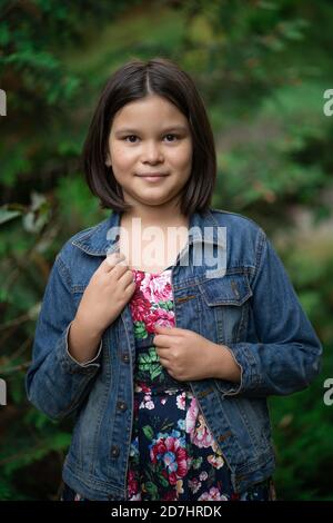 Cute little girl wearing plaid dress and denim jacket posing against backdrop of green nature Stock Photo