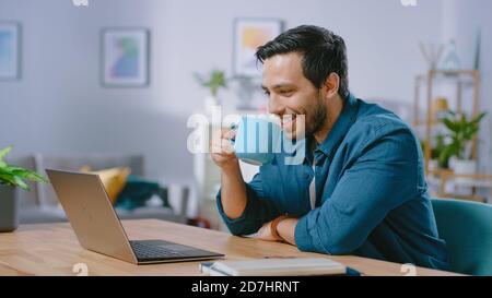Portrait of Smiling Young Man Working on a Laptop from Home and Drinking Coffee From the Mug and is Satisfied with His Work. Cozy Modern Flat done in Stock Photo