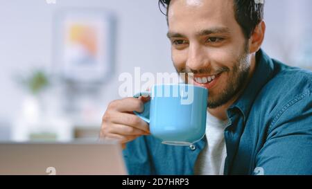 Portrait of Focused Young Man Working on a Laptop from Home, He Drinks Beverage From the Mug and is Satisfied with His Work. Cozy Modern Flat done in Stock Photo