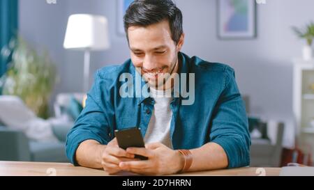 Handsome Smiling Man Uses Smartphone while Sitting at the Desk of His Living Room. Guy Browsing in Internet, Using Social Networks, Relaxing at Home. Stock Photo