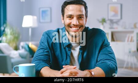 Portrait of Handsome Man Sitting at Desk in His Living Room and Charmingly Smiling. Medium Shot. Stock Photo