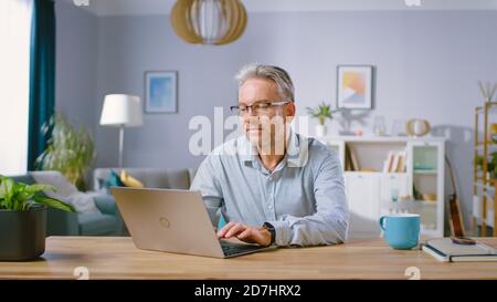 Handsome Middle Aged Man Uses Laptop Computer While Sitting at His Desk in the Cozy and Stylish Living Room. Happy Successful Man Works on Computer Stock Photo