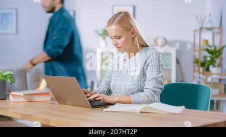 Beautiful Woman Works on a Laptop at Home, Her Boyfriend Walks by in the Background. Professional Female Freelancer Using Computer. Stock Photo
