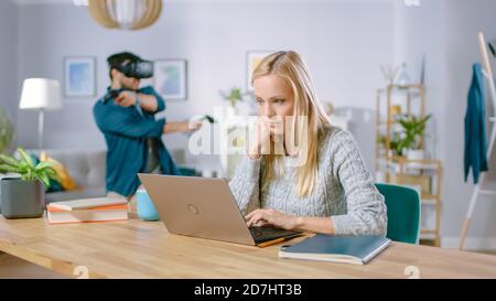 Beautiful Concentrated Woman Works on a Laptop at Her Desk, Behind Her Boyfriend Wearing Virtual Reality Headset Plays in Video Game with Controllers Stock Photo