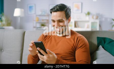 Happy Young Man Uses Smartphone while Sitting on a Sofa at Home. Man Browses Through Internet, Watches Videos and Uses Social Networks at Home. Stock Photo