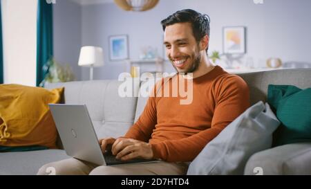 Cheerful Young Man at Home Sitting on a Sofa Holds Laptop on His Lap, Browses Through the Internet, Social Networks, Does e-Shopping. Stock Photo