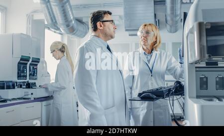 Two Research Scientists Talking about Programming Medical Equipment for Tests. Team of Professionals Doing Pharmaceutical Research in Modern Stock Photo