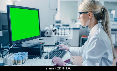 Female Research Scientist Sits at His Workplace in Laboratory, Uses Green Mock-up Screen Personal Computer. In the Background Genetics, Pharmaceutical Stock Photo