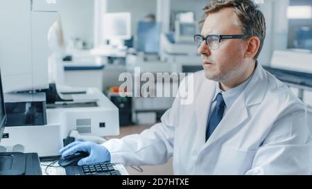 Male Research Scientist Sits at His Workplace in Laboratory, Uses Personal Computer. I the Background Genetics, Pharmaceutical Research Centre with Stock Photo