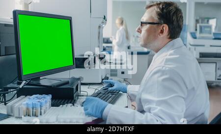 Male Research Scientist Sits at His Workplace in Laboratory, Uses Green Mock-up Screen Personal Computer. I the Background Genetics, Pharmaceutical Stock Photo