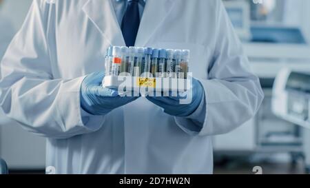 Close-up Shot of a Male Research Scientist Stands in Laboratory and Hold Tray of Test Tubes Filled with Samples. In the Background Working Laboratory Stock Photo