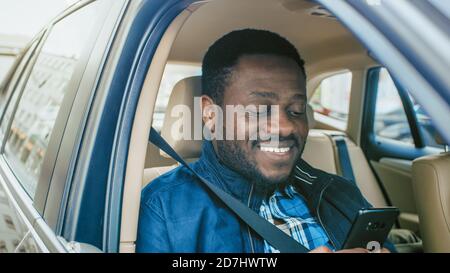 Handsome Entrepreneur Traveling on a Passenger Seat of a Car, Uses Smartphone and Smiles. Camera Shot from Outside the Vehicle. Stock Photo