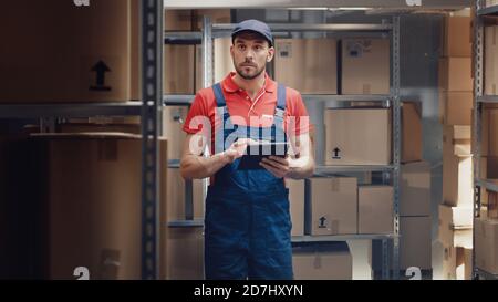 Warehouse Worker Uses Digital Tablet For Checking Stock, On the Shelves Standing Cardboard Boxes. Stock Photo