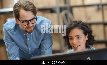 Male and Female Warehouse Inventory Managers Talking, Using Personal Computer and Checking Stock. In the Background Rows of Shelves Full of Cardboard Stock Photo