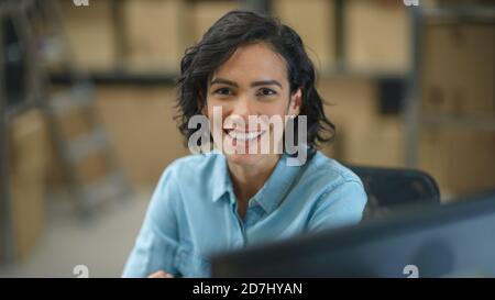 Female Inventory Manager Works Sitting at Her Desk and Smiling at the Camera. In the Background Warehouse Storeroom with Shelves Full of Cardboard Box Stock Photo