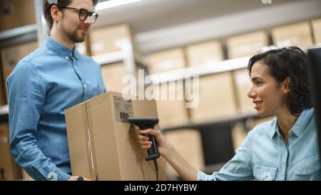 Female Inventory Manager Scans Cardboard Box and Digital Tablet with Barcode Scanner, Male Worker Holds Package. In the Background Rows of Cardboard Stock Photo