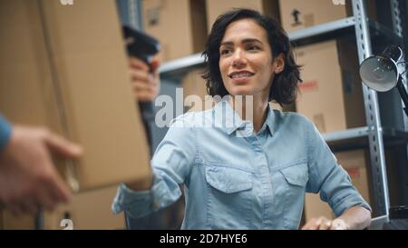 Female Inventory Manager Scans Cardboard Box and with Barcode Scanner, Male Worker Holds Package. In the Background Rows of Cardboard Boxes with Stock Photo