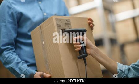 Female Inventory Manager Scans Cardboard Box and with Barcode Scanner, Male Worker Holds Package. In the Background Rows of Cardboard Boxes with Stock Photo