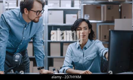 Male and Female Warehouse Inventory Managers Talking, Using Personal Computer and Checking Stock. Female Pointing at Screen. In the Background Rows of Stock Photo