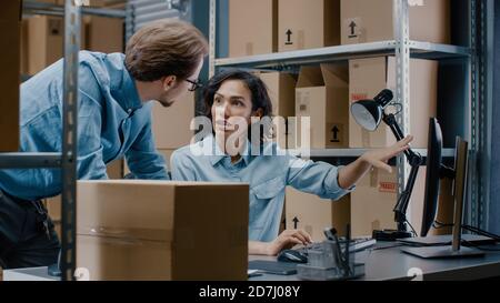 Male and Female Warehouse Inventory Managers Talking, Using Personal Computer and Checking Stock. In the Background Rows of Shelves Full of Cardboard Stock Photo