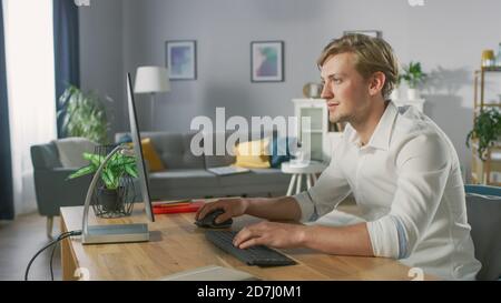 Handsome Young Designer Works On Personal Computer Sitting at His Desk. In the Background Stylish Home Decor. Stock Photo