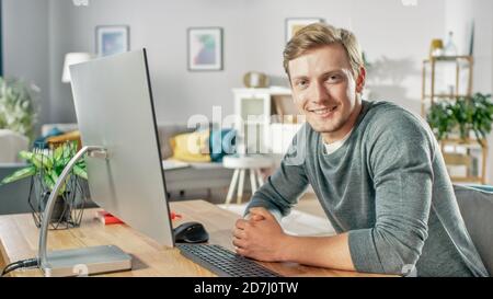 Portrait of the Focused Young Man Working on a Personal Computer while Sitting at His Desk, Smiling and Looking at the Camera. In the Background Cozy Stock Photo