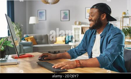 Portrait of the Smiling Stylish African American Working on a Personal Computer while Sitting at His Desk at Home. Stock Photo