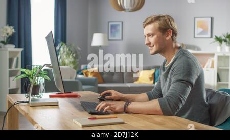 Portrait of the Handsome Man Working on Personal Computer while Sitting at His Desk. In the Background Stylish Cozy Living Room. Young Man Playing Stock Photo