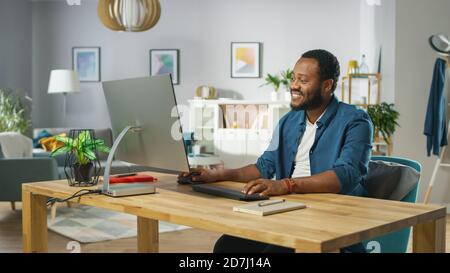 Happy Young Man Takes Place at His Desk and Starts Working on a Personal Computer. In the Background Cozy Living Room. Stock Photo