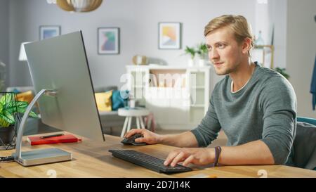 Portrait of the Focused Young Man Working on a Personal Computer while Sitting at His Desk. In the Background Cozy Living Room. Stock Photo