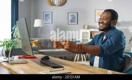 Portrait of the Handsome African American Man Pointing at the Screen with Two Fingers while Sitting at His Workplace. Young Man Having Fun at Home. Stock Photo