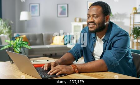 Handsome Smiling Black Man Using Laptop While Sitting at the Desk of His Cozy Living Room. Stock Photo