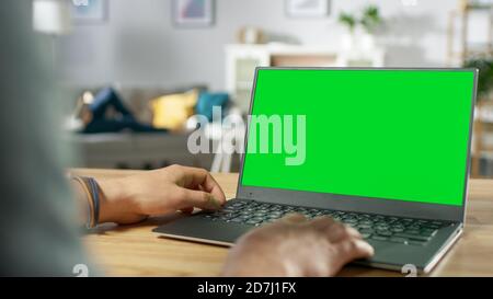 Over the Shoulder Shot of Mans Hands Using Laptop with Green Mock-up Screen. In the Background Cozy Living Room with Woman Relaxing on a Sofa. Stock Photo