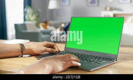 Over the Shoulder Shot of Mans Hands Typing on a Laptop with Green Mock-up Screen. In the Background Cozy Living Room with Woman Relaxing on a Sofa. Stock Photo