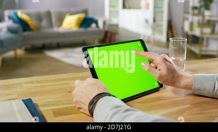 Man Holding and Using Hand Gestures on Green Mock-up Screen Digital Tablet Computer While Sitting at the Desk. Man Buying stuff or Browsing Through Stock Photo