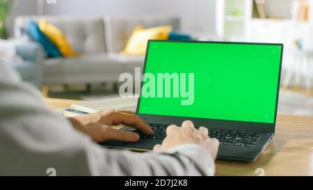 Close-up of a Man Uses Laptop with Green Mock-up Screen While Sitting at the Desk in His Cozy Living Room. Stock Photo