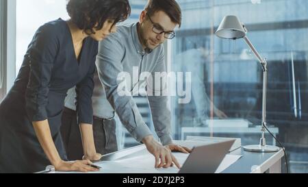 Two Young Business Entrepreneurs Leaning on a Desk, Working with Documents for a New Startup Project. Businesspeople Using Laptop in the Office with Stock Photo