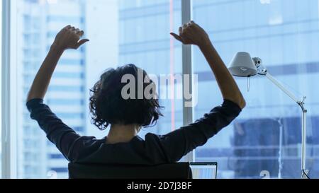 Back View Shot of the Beautiful Businesswoman Sitting at Her Office Desk, Raising Arms in a Celebration of a Successful Job Promotion. Stock Photo