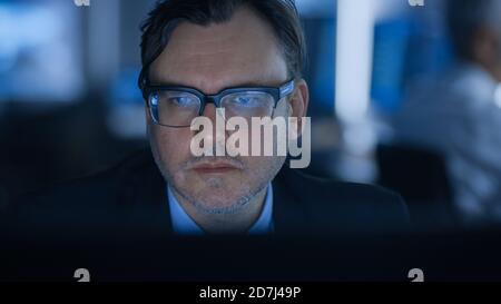 Portrait of the Handsome Expert wearing Glasses Working on a Computer. In the Background Out of Focus Professionals Working. Stock Photo