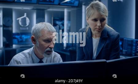 Female Manager Talks with Professional Operator Working on a Personal Computer in the System Monitoring Room. High Profile Specialists Finding Stock Photo