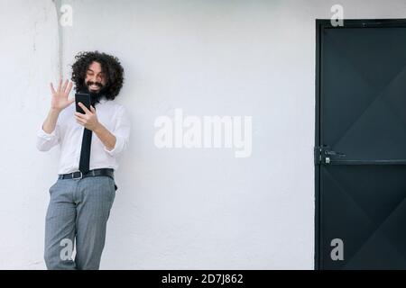Businessman waving hand to video call on phone while standing against wall Stock Photo