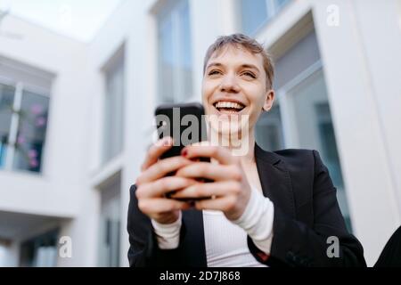 Close-up of cheerful trans young man using mobile phone while sitting against building Stock Photo