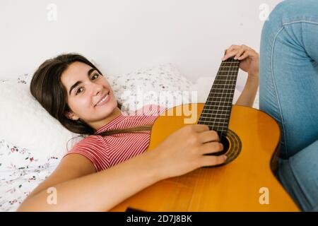 Young woman lying down while playing guitar in bedroom Stock Photo
