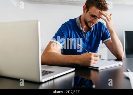 Smiling handsome man reading from book while sitting with laptop at home Stock Photo