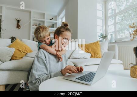 Woman working on laptop while boy hugging her from behind at home Stock Photo