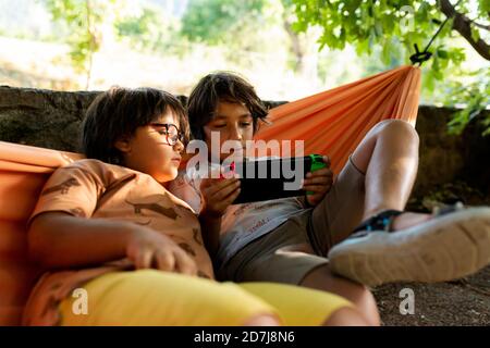 Siblings playing video games while lying on hammock at backyard Stock Photo