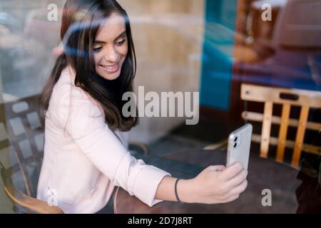 Smiling young female entrepreneur taking selfie through smart phone in cafe Stock Photo